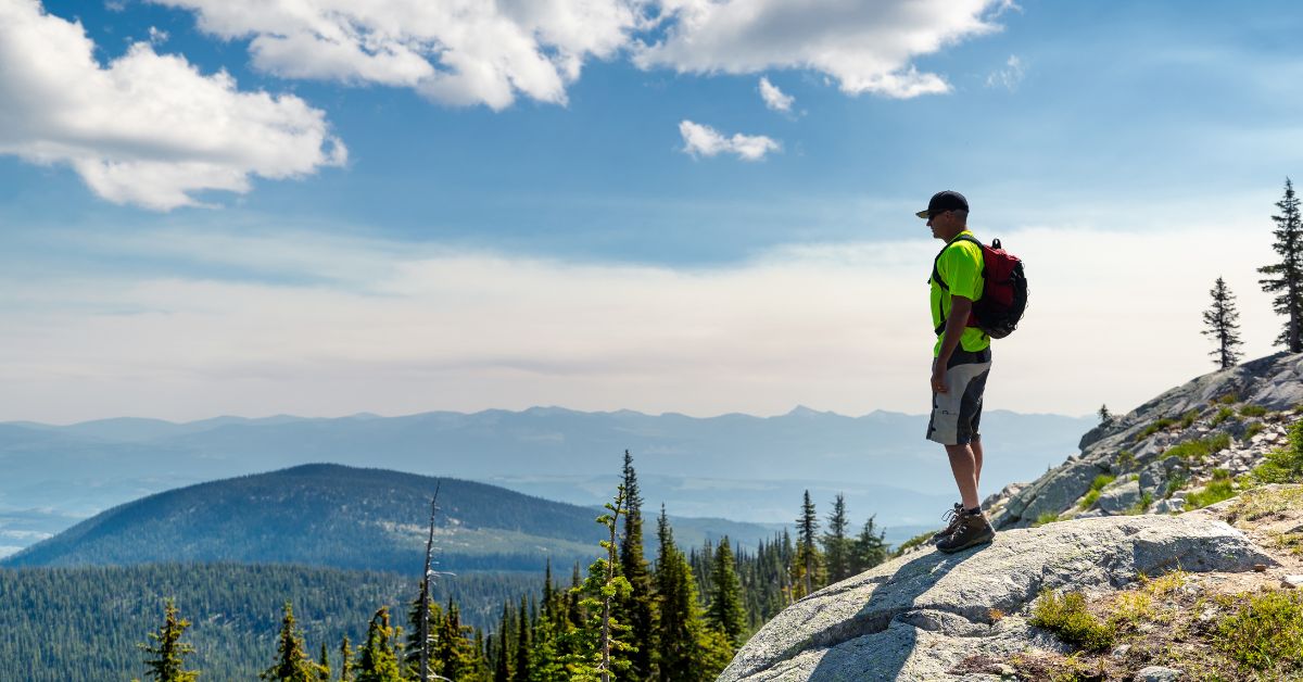 A man with a small backpack stands atop a mountain and looks out at his surroundings