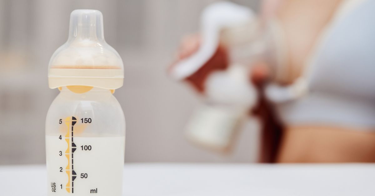 A baby bottle full of milk with a woman in the background breast pumping
